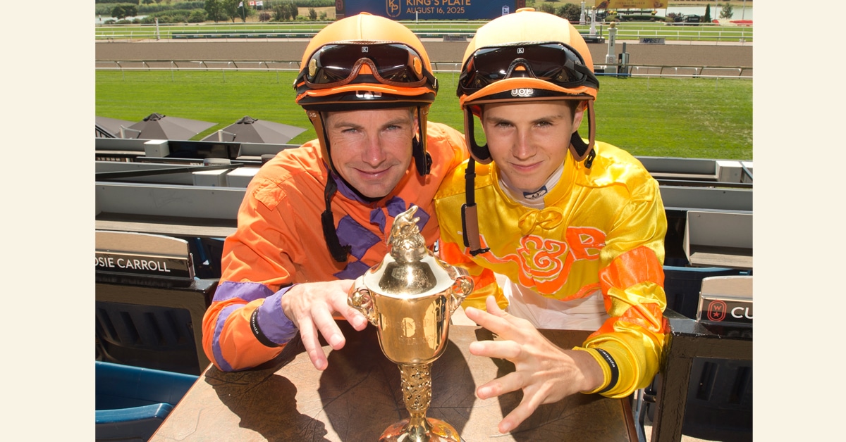 Two jockeys reaching for a trophy on a table.
