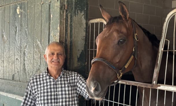 A man standing with a bay horse in its stall at the track.
