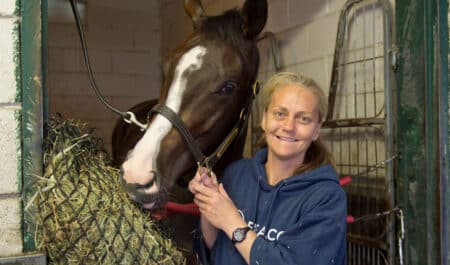 A blonde woman holding a bay horse in a stall at the track.