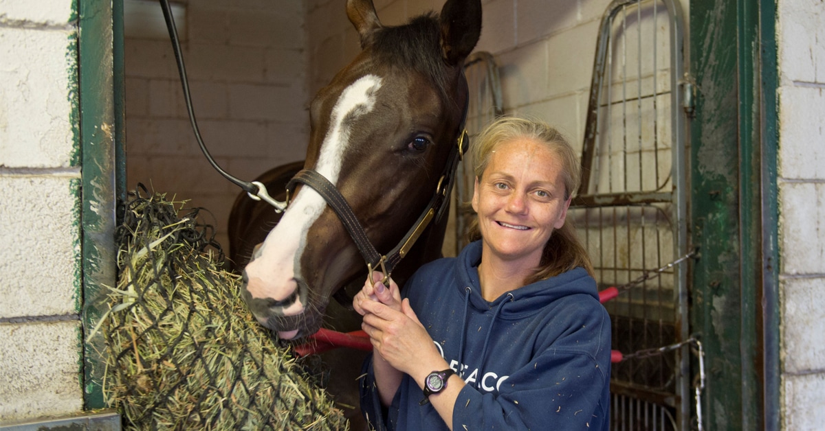 A blonde woman holding a bay horse in a stall at the track.