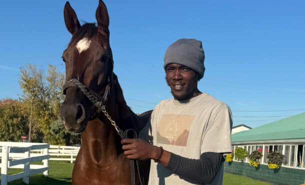 A man holding a bay horse on the backstretch.