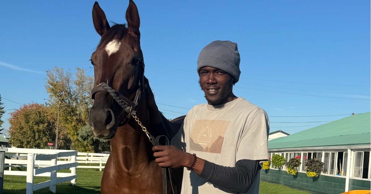 A man holding a bay horse on the backstretch.
