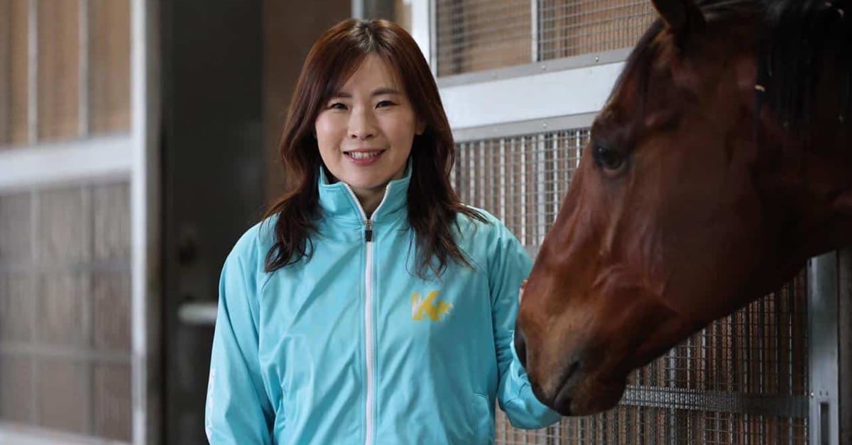 A smiling woman holding a bay horse in a racing stable.