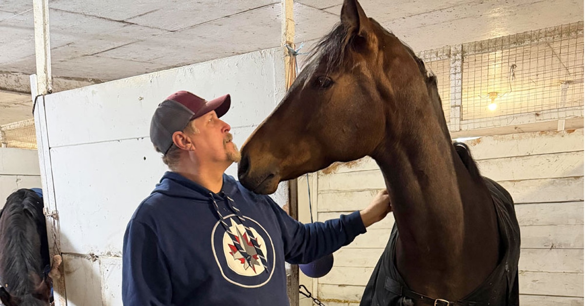 A man patting a bay horse in a stall at the track.