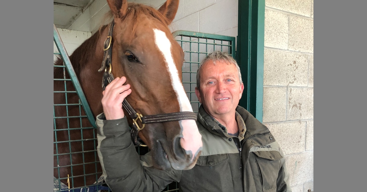 A man standing with a chestnut horse in a backstretch stall.