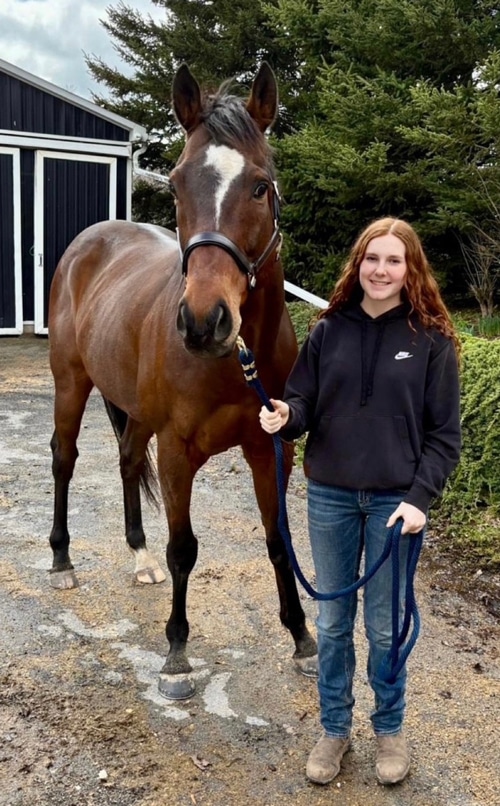 A young woman holding a bay horse in front of a barn.